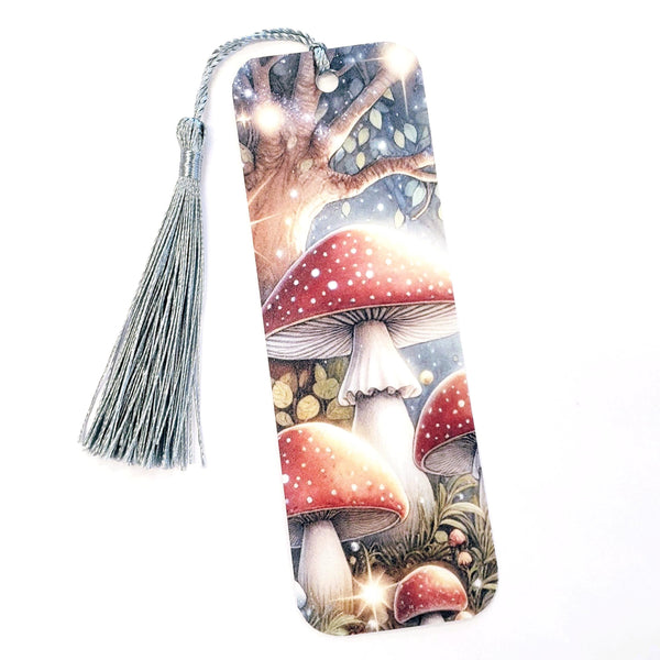 A bookmark with a silver tassel. The bookmark features a watercolor illustration of red and white mushrooms in a forest setting. The mushrooms have white stems and red caps with white spots. The background is a blurred image of a forest with green leaves and a blue sky. The bookmark is a great gift for mushroom lovers and nature enthusiasts.