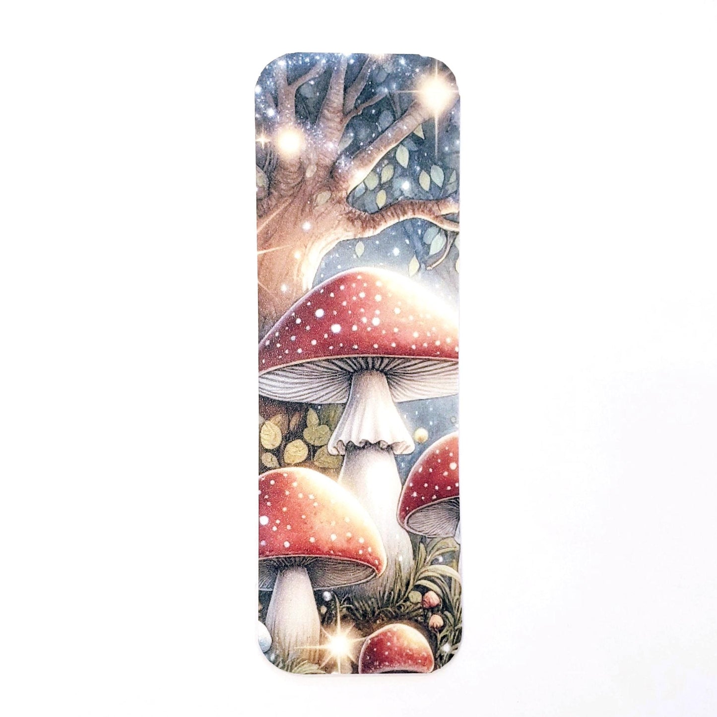 A bookmark with a whimsical illustration of red and white mushrooms in a forest setting. The mushrooms have white stems and red caps with white spots. The background features a tree with a blue sky and twinkling stars. The bookmark is a great way to mark your place in a book and add a touch of magic to your reading experience.