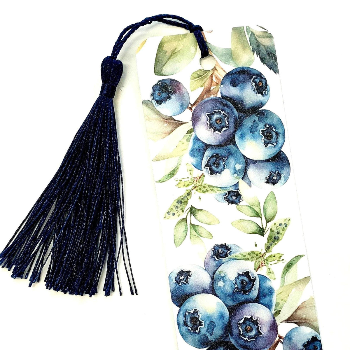A white bookmark with a blue tassel. The bookmark features a watercolor illustration of blueberries on a branch with green leaves.