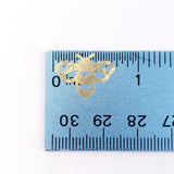 A gold metallic bee sticker measuring approximately 0.75 inches in width. The bee is shown against a blue ruler with black markings.