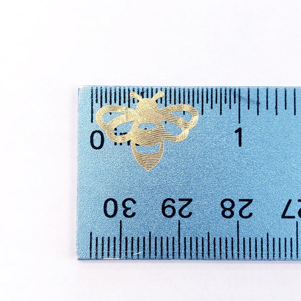 A gold metallic bee sticker measuring approximately 0.75 inches in width. The bee is shown against a blue ruler with black markings.