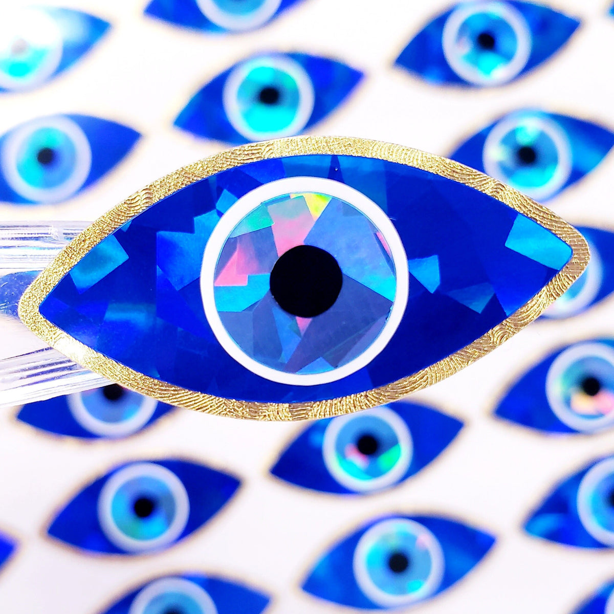 A close-up of a blue evil eye sticker with a gold rim. The eye is iridescent and has a white center with a black pupil. Th
